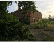 Backsteingebäude mit roten Dachziegeln, umgeben von dichtem grünen Bewuchs und einer verwilderten Betonfläche im Vordergrund; im Hintergrund sind hohe Bäume und ein teils bewölkter Himmel zu sehen.