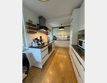 Moderne Küche mit weißer Hochglanz-Einrichtung, eingebauten Elektrogeräten und schwarzen Arbeitsplatten. Der Raum hat einen Holzfußboden, offene Regale mit Gewürzen und ist durch eine große Fensterfront gut beleuchtet.