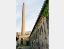 Backsteingebäude mit einem hohen Schornstein, umgeben von einem Innenhof mit gepflastertem Boden; rechts ist eine mit Efeu überwucherte Wand sichtbar. Im Hintergrund befinden sich Gebäude mit moderneren Fenstern.