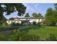 Ein zweistöckiges Gebäude mit heller Fassade und roten Backsteinakzenten, umgeben von grüner Vegetation und Bäumen. Ein gepflasterter Weg führt durch einen Rasenbereich zur Eingangstür.