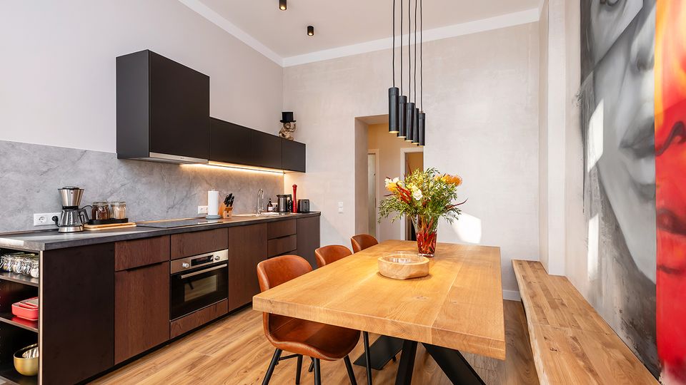 Moderne Wohnküche mit dunkler, griffsloser Einbauzeile und grauer Rückwand, Holzboden sowie langem Holzesstisch mit Lederstühlen. Über dem Tisch hängen schwarze Pendelleuchten; an der rechten Wand befindet sich ein großformatiges Wandbild.