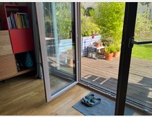 Ein offenes Glaselement einer Terrassentür führt von einem Innenraum mit Holzboden und Regal zu einer Holzterrasse mit Blumentöpfen und grüner Bepflanzung. Im Vordergrund liegt eine Fußmatte mit Sandalen darauf.