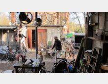 Eine Werkstatt oder Hinterhofszene mit einer chaotischen Anordnung von Fahrrädern, Werkzeugen und recycelbaren Gegenständen. Im Hintergrund ein Mann in dunkler Kleidung, umgeben von Bäumen, einer Ziegelwand und einem parkenden grünen Transporter.