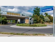 Kreisverkehr unter einer Brücke mit bepflanzter Mittelinsel und einem modernen Kunstwerk aus Metall. Im Hintergrund sind Bäume und ein Straßenschild mit der Aufschrift „Ostverbinder“ sichtbar.