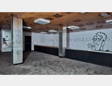 Leerer Innenraum eines verlassenen Laden- oder Gastronomiebereichs mit langer Theke entlang der Rückwand, Metallstützen und abgehängter Decke mit rechteckigen Leuchten. Weiße Wandpaneele mit kleinen Öffnungen und Graffiti, dunkler Fliesenboden.