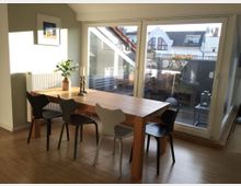 Essbereich in einem modernen Innenraum mit einem Holztisch und sechs unterschiedlich gestalteten Stühlen auf einem Parkettboden. Im Hintergrund große Fensterfront mit Blick auf eine Terrasse und umliegende Gebäude.