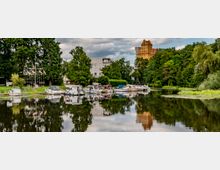 Bootsanleger an einem Fluss mit mehreren festgemachten Motorbooten, umgeben von grünen Bäumen. Im Hintergrund ist ein modernes Gebäude sowie ein markantes, orangefarbenes Haus zu sehen.