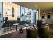 Moderner Veranstaltungsraum in einem ehemaligen Kirchengebäude mit Stuhlreihen, einer Bühne mit Ledersesseln und einem Rednerpult. Im Hintergrund eine beleuchtete Wand mit einem großen Kreuz und einem bunten Glasfenster, flankiert von hohen Fenstern und Galerien.