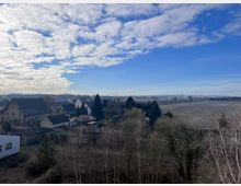 Blick auf ein ländliches Dorf mit vereinzelten Häusern und Gärten, umgeben von kahlen Bäumen und Feldern unter einem teils wolkigen blauen Himmel. Im Hintergrund erstreckt sich eine flache Landschaft mit Bäumen und einer entfernten Stromleitung.