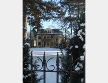 Gelbes zweistöckiges Herrenhaus im historischen Stil, umgeben von verschneiten Bäumen und einem umzäunten Garten, fotografiert im Winter bei klarem Himmel. Im Vordergrund ist ein mit Schnee bedeckter schwarzer Metallzaun sichtbar.