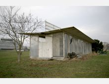 Ein einfaches, rechteckiges Gebäude mit flachem Dach aus Metall und Glaswänden, von denen einige beschädigt sind. Im Vordergrund stehen ein kahler Baum und ein Stapel Rohre, das Gebäude befindet sich auf einer Wiese inmitten einer offenen Umgebung mit anderen Gebäuden im Hintergrund.