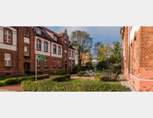 Backstein-Wohngebäude mit weißen Fassadenelementen und Satteldächern, umgeben von gepflegten Hecken und Bäumen. Im Vordergrund befinden sich ein gepflasterter Weg, Fahrradständer mit abgestellten Fahrrädern und ein Wegweiser mit Straßennamen.