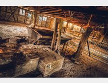 Innenraum eines historischen Industriegebäudes mit einer alten Lore und metallenen Stützelementen. Der Raum ist aus Stein und Holz gebaut, mit Pflasterboden, Holzbalken und mehreren Fenstern, die diffuse Beleuchtung einlassen.
