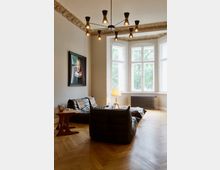 Wohnzimmer mit Parkettboden, großen weißen Sprossenfenstern und einer dekorativen Stuckdecke. Im Raum befinden sich schwarze Ledersessel, ein kleiner Beistelltisch, eine Stehlampe und ein Wandbild.