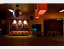 Innenraum eines Clubs oder Veranstaltungsraums mit einer Disco-Kugel an der Decke, mehreren orange gepolsterten Stühlen entlang der Wand und gedämpfter Beleuchtung. Die Wände sind in warmen Farbtönen gehalten, und ein großes Wandbild zeigt eine historische Szene.