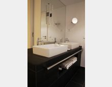 Moderne Badezimmeransicht mit schwarzer Waschtischkonsole, zwei weißen rechteckigen Waschbecken und einem großen Wandspiegel. Darunter liegen sauber gefaltete Handtücher in einem offenen Regal, mit minimalistischem Design und einer runden Wandleuchte an der weißen Wand.