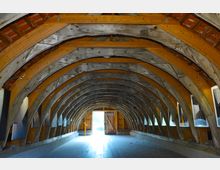 Holzstrukturierte Halle mit gewölbtem Dach aus dicken Holzträgern und modernen unterstützenden Elementen. Die Wände verfügen über eingelassene Fenster und Poster, während eine offene Tür im Hintergrund Tageslicht einlässt.