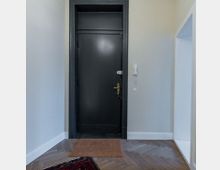 Ein schlichter Flur mit heller Wandgestaltung, einer schwarzen Eingangstür, Parkettboden im Fischgrätmuster, einer Fußmatte und einem Wandtelefon mit Gegensprechanlage an der rechten Seite.