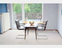 Ein kleiner Raum mit einem rechteckigen Holztisch und zwei schwarzen Stühlen auf einem hellen Teppichboden. Im Hintergrund ist eine große Fensterfront mit Blick auf einen Balkon, eine Holzzaunbegrenzung und grüne Bäume zu sehen.