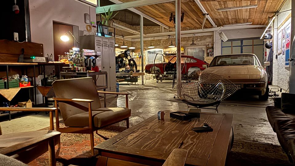 Große Werkstatt- oder Garagenhalle mit Holzdecke und Stahlträgern, in der mehrere Autos stehen und Werkbänke sowie Geräte im Hintergrund zu sehen sind. Im Vordergrund befindet sich ein Lounge-Bereich mit Sesseln, Couchtisch und Teppich auf Betonboden, seitlich hängen Poster und Lampen an den Wänden.
