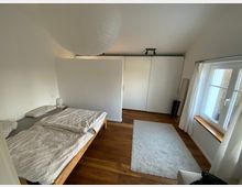 Schlafzimmer mit minimalistischem Design: Ein Doppelbett mit heller Bettwäsche steht auf einem Holzboden, daneben ein kleiner Nachttisch mit Lampe. Im Hintergrund befinden sich ein weißer Wandschrank, ein Teppich und ein Spiegel an der Wand. Ein großes Fenster mit weißen Vorhängen lässt Tageslicht herein.