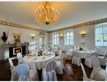 Eleganter Veranstaltungsraum mit runden Tischen, weißen Tischtüchern und Stuhlhussen, dekoriert mit Kerzenhaltern und gefalteten Servietten. Im Hintergrund befinden sich ein Kamin mit Wappen und ein großer funkelnder Kronleuchter, eingerahmt von weißen Wänden mit goldenen Verzierungen und Fenstern, die Tageslicht hereinlassen.