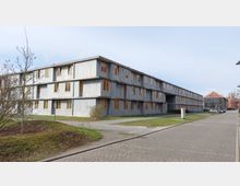 Flaches, graues Betongebäude mit drei Stockwerken und zahlreichen Fenstern mit orangefarbenen Rahmen, umgeben von Grünflächen und einem gepflasterten Gehweg; im Hintergrund ist ein weiteres modernes Gebäude mit rotem Dach zu sehen.