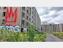 Mehrstöckige verlassene Wohngebäude mit zerbrochenen Fenstern und Graffiti auf den Wänden, umgeben von verwilderter Vegetation und einem asphaltierten Weg im Vordergrund. Im Hintergrund sind moderne Wohnhochhäuser und bewölkter Himmel zu sehen.