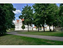 Historisches Gebäude mit weißer Fassade und roten Giebel­dächern, umgeben von Bäumen und einer Grünfläche mit einem geschwungenen Spazierweg im Vordergrund. Im Hintergrund sind weitere Gebäude mit ähnlicher Architektur zu sehen.