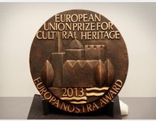 Bronzene Auszeichnung mit der Inschrift „European Union Prize for Cultural Heritage - Europa Nostra Award 2013“, verziert mit einer Reliefdarstellung von Gebäuden, Bäumen und Wellen, auf schwarzem Sockel präsentiert.