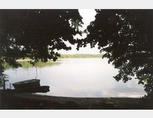 Ein See mit ruhiger Wasseroberfläche, umrahmt von überhängenden Baumästen im Vordergrund. Links im Bild liegt ein kleines Ruderboot am sandigen Ufer.