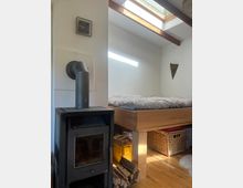 Kleiner Innenraum mit einem Holzofen auf der linken Seite, einem erhöhten Bettgestell aus Holz auf der rechten Seite und einem Dachfenster, das Tageslicht hereinlässt. Unter dem Bett befinden sich Körbe und Kisten zur Aufbewahrung, daneben liegt gestapeltes Brennholz.