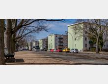 Straßenszene in einem Wohngebiet mit mehrstöckigen, hellgrün gestrichenen Wohnhäusern. Im Vordergrund führen eine Bank, große, laublose Bäume und geparkte Autos entlang der Straße.