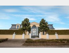 Gelbes Einfamilienhaus mit symmetrischer Fassade, schwarzem Mansardendach und zentralem Eingangsbereich mit Glasportal, flankiert von zwei hohen Bäumen. Vor dem Haus befindet sich eine niedrige Mauer mit Hecke und dekorativen weißen Säulen sowie ein schmiedeeisernes Tor.