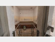 Kleiner, karg ausgestatteter Raum mit abgenutzten Wänden und einer erhöhten Holzplattform. Im Vordergrund befindet sich ein Metallgeländer, dahinter ein grüner Eimer auf dem Boden.