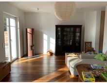 Helles Wohnzimmer mit Holzboden, einer großen Glastür auf der linken Seite, die Tageslicht hereinlässt, und einer zentral platzierten Papierlampenschirm-Deckenleuchte. Es gibt einen braunen Kamin, eine dunkle Bücherwand, ein Sofa mit Spielzeug darauf und einen Holztisch im Vordergrund mit Gesellschaftsspielen.