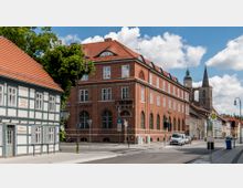 Straßenszene mit einem historischen Fachwerkhaus links und einem großen roten Backsteingebäude im Zentrum, das als Sparkasse gekennzeichnet ist. Im Hintergrund ist eine Kirche mit spitzem Kirchturm und breiter Turmbasis zu sehen, daneben stehen geparkte Autos und Straßenlaternen.