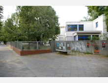 Außenansicht eines modernistischen Gebäudes mit weißen Wänden und blauen Fenstern, umgeben von einem Zaun und Bäumen. Im Vordergrund befindet sich ein gepflasterter Gehweg, eine Infotafel sowie eine Mauer mit Plakaten und Graffiti.
