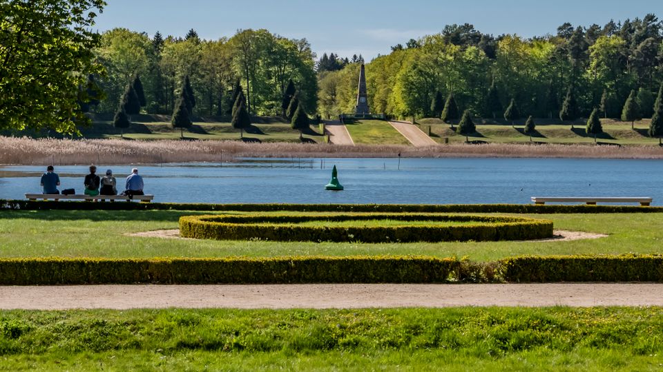 Lakeside park with manicured lawns, low boxwood hedges in geometric beds, and benches along the water’s edge. Across the lake, a tree-lined slope leads to a narrow obelisk-like monument framed by forest.