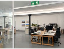 Heller Großraumbüro- oder Atelierraum mit grauem Betonboden, weißen Wänden und langen Deckenleuchten, in der Mitte von einer schwarzen Stützsäule getragen. Mehrere Schreibtische mit Monitoren und Arbeitslampen stehen neben Regalen, Pflanzen und einer Kleiderstange; links ist eine Glaswand und oben ein grünes Notausgangsschild.