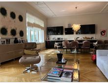 Wohnbereich einer modernen Wohnung mit offener Küche. Der Raum hat einen Parkettboden, helle Wände, große Fenster mit Rollos, eine Sitzecke mit einem braunen Sessel und Glastisch sowie eine schwarze Küchenzeile, vor der ein Esstisch mit rosafarbenen Stühlen steht.