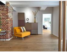 Wohnzimmer mit modernem Interieur, bestehend aus einem gelben Sessel, einer minimalistisch gestalteten Kommode und Dekoration wie einer Vase und einer Lampe. Links ist eine freiliegende Backsteinwand zu sehen, im Hintergrund ein weiterer Raum mit einem Gemälde an der Wand und einem Sessel.