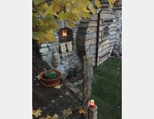 Steinerne Gebäudeecke mit kleinem, vergittertem Fenster und warmem Licht im Inneren, daneben ein holzverkleideter Anbau mit kleiner Tür und Fenster. Im Vordergrund Gartenbereich mit Pflanzschale, Drahtzaun, Holzpfosten und herbstlichen Ahornblättern.