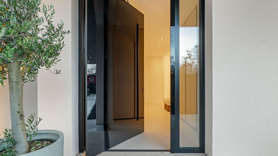 Eingangsbereich eines modernen Wohnhauses mit geöffneter, dunkler Haustür und seitlichen Glasflächen in schwarzem Rahmen. Vor der Tür stehen große Steinfliesen und ein Pflanzkübel mit kleinem Baum; innen ist ein heller Flur mit Deckenspots zu sehen.