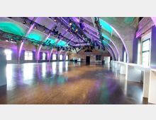 Großer Veranstaltungsraum mit einem hohen, gewölbten Dach aus Beton und modernen Lichtinstallationen in Lila und Grün. Die Wände kombinieren Ziegelstein und Glasfenster, während der Raum mit glänzendem Boden und weißen Stehtischen ausgestattet ist.