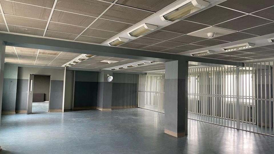 Großer, leerer Innenraum mit grauen Wänden und blau-grauem Boden, getragen von Stützpfeilern und einer abgehängten Decke mit langen Leuchtstofflampen. Rechts befindet sich ein Bereich mit Metallgitter-Trennwänden vor Fensterfronten, links sind mehrere offene Türdurchgänge zu weiteren Räumen.