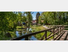 Holzbrücke über einen schmalen, baumbestandenen Fluss in ländlicher Umgebung mit Wiesen, einem Traktor und kleinen Gebäuden im Hintergrund. Ein asphaltierter Weg führt entlang des Flusses, auf dem zwei Personen mit Fahrrädern zu sehen sind.