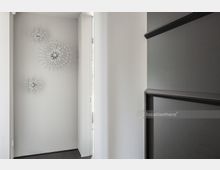 Moderner, minimalistischer Innenraum mit weißer Wand, an der drei silberne, sternförmige Wanddekorationen angebracht sind. Rechts ist ein dunkles Regal sichtbar, links ein schmaler Türrahmen und ein Fenster lässt Tageslicht herein.