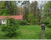 Ein Garten am Waldrand mit einer kleinen, weiß verputzten Hütte mit rotem Ziegeldach. Im Vordergrund stehen grüne Sträucher und Wiesen, während im Hintergrund ein dichter Laub- und Nadelwald sowie eine überdachte Holzschaukel zu sehen ist.