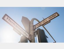 Straßenschilder an einer Kreuzung, die die "Hermannstraße" und "Flughafenstraße" anzeigen, vor einem klaren blauen Himmel; im Vordergrund sind Teile einer Ampelanlage zu sehen.